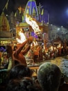A vibrant religious ceremony taking place by a river at night. A man in traditional attire holds up a burning lamp, surrounded by other participants. The area is bustling with people, with ornate structures and flags in the background. The scene is lit by the flames of the lamps, creating a warm glow.