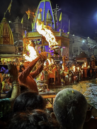 A vibrant religious ceremony taking place by a river at night. A man in traditional attire holds up a burning lamp, surrounded by other participants. The area is bustling with people, with ornate structures and flags in the background. The scene is lit by the flames of the lamps, creating a warm glow.
