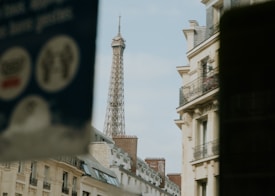 A street view featuring the Eiffel Tower in the background, partially obscured by residential buildings. The scene includes ornate balconies and classic Parisian architectural elements. A blurred sign or poster is partially visible in the foreground, adding an urban feel.