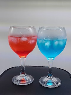 Two glass goblets filled with colorful drinks, one red and the other blue, on a dark surface. The goblets contain ice cubes, suggesting the beverages are cold and refreshing.