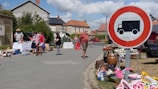 A street market scene with people browsing tables filled with various items for sale. A large circular road sign with a truck symbol is prominently displayed. In the background, houses with red roofs and some greenery are visible under a partly cloudy sky.