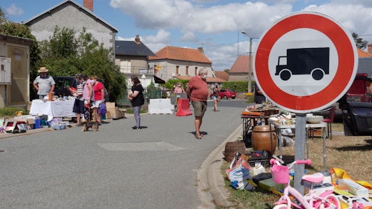 A street market scene with people browsing tables filled with various items for sale. A large circular road sign with a truck symbol is prominently displayed. In the background, houses with red roofs and some greenery are visible under a partly cloudy sky.