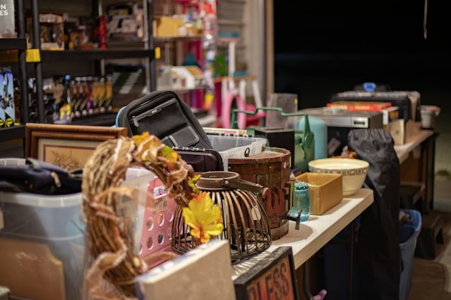 A table is filled with a variety of items, including a wicker wreath with yellow flowers, a black bag, several baskets, a framed picture, a bowl, and a blue jar. Shelves in the background hold more assorted goods.