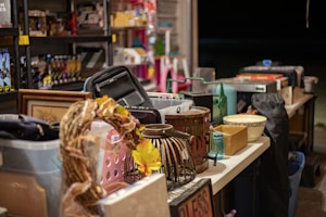 A table is filled with a variety of items, including a wicker wreath with yellow flowers, a black bag, several baskets, a framed picture, a bowl, and a blue jar. Shelves in the background hold more assorted goods.