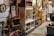A cluttered antique store or flea market display with various vintage items on shelves and tables. There are old bottles, a fan, books, decorative plates, vintage signs, and a duck figurine. The setting is a dimly lit room with multiple wooden and metal shelving units filled with collectables and artifacts, giving a rustic and nostalgic feel.