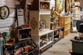 A cluttered antique store or flea market display with various vintage items on shelves and tables. There are old bottles, a fan, books, decorative plates, vintage signs, and a duck figurine. The setting is a dimly lit room with multiple wooden and metal shelving units filled with collectables and artifacts, giving a rustic and nostalgic feel.