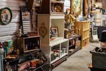 A cluttered antique store or flea market display with various vintage items on shelves and tables. There are old bottles, a fan, books, decorative plates, vintage signs, and a duck figurine. The setting is a dimly lit room with multiple wooden and metal shelving units filled with collectables and artifacts, giving a rustic and nostalgic feel.