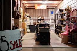 A well-lit storage room filled with a variety of items organized on shelves. A prominent sign in the foreground advertises a sale for items priced at $5 or less. The space is cluttered with various household goods, including furniture, toys, and decorations, creating a busy and eclectic atmosphere.
