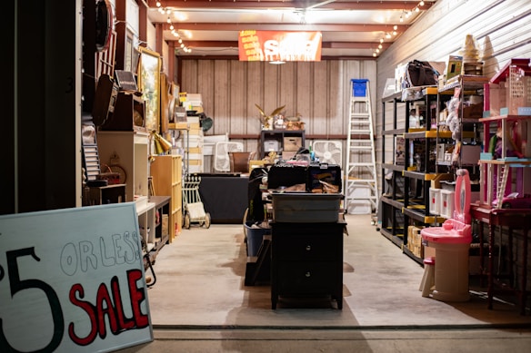 A well-lit storage room filled with a variety of items organized on shelves. A prominent sign in the foreground advertises a sale for items priced at $5 or less. The space is cluttered with various household goods, including furniture, toys, and decorations, creating a busy and eclectic atmosphere.