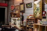 A room filled with an eclectic collection of vintage and retro items. Various framed artworks and memorabilia are displayed on the walls. A collection of small objects and antiques are arranged on shelves and cabinets, with decorative elements like glass bottles, figurines, and a lamp. The ambiance is nostalgic, evoking a sense of history and charm.