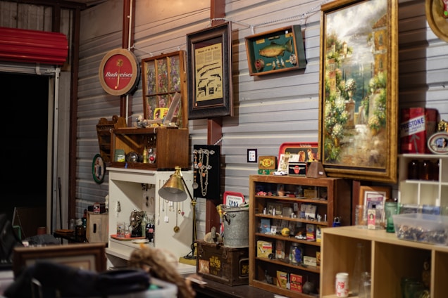 A room filled with an eclectic collection of vintage and retro items. Various framed artworks and memorabilia are displayed on the walls. A collection of small objects and antiques are arranged on shelves and cabinets, with decorative elements like glass bottles, figurines, and a lamp. The ambiance is nostalgic, evoking a sense of history and charm.