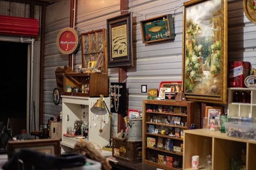 A room filled with an eclectic collection of vintage and retro items. Various framed artworks and memorabilia are displayed on the walls. A collection of small objects and antiques are arranged on shelves and cabinets, with decorative elements like glass bottles, figurines, and a lamp. The ambiance is nostalgic, evoking a sense of history and charm.