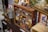 A variety of vintage and collectible items are displayed on wood shelves. There are several metal tins, boxes, old toy cars, and assorted memorabilia. The cabinet appears to be in a room with other collectibles, including a rustic metal container and framed artwork.