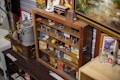 A variety of vintage and collectible items are displayed on wood shelves. There are several metal tins, boxes, old toy cars, and assorted memorabilia. The cabinet appears to be in a room with other collectibles, including a rustic metal container and framed artwork.