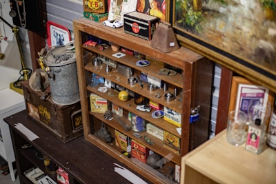 A variety of vintage and collectible items are displayed on wood shelves. There are several metal tins, boxes, old toy cars, and assorted memorabilia. The cabinet appears to be in a room with other collectibles, including a rustic metal container and framed artwork.