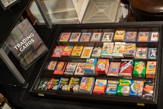 A display case filled with various trading cards, including baseball and football cards, with vibrant packaging. A sign labeled 'Trading Cards' is positioned nearby, indicating the collection available for viewing.