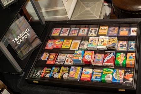 A display case filled with various trading cards, including baseball and football cards, with vibrant packaging. A sign labeled 'Trading Cards' is positioned nearby, indicating the collection available for viewing.