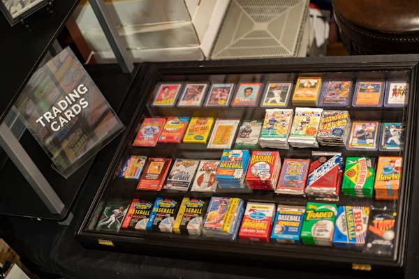 A display case filled with various trading cards, including baseball and football cards, with vibrant packaging. A sign labeled 'Trading Cards' is positioned nearby, indicating the collection available for viewing.