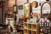 A variety of vintage items are displayed on shelves in a cluttered space, including framed pictures, signs, and glassware. There are decorative items such as old signs, a lamp, and a mirror with a wooden frame. Shelves are filled with assorted collectibles, including small jars, boxes, and figurines.