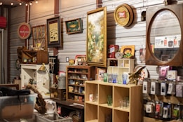 A variety of vintage items are displayed on shelves in a cluttered space, including framed pictures, signs, and glassware. There are decorative items such as old signs, a lamp, and a mirror with a wooden frame. Shelves are filled with assorted collectibles, including small jars, boxes, and figurines.