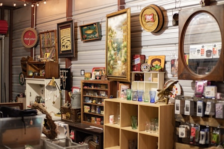 A variety of vintage items are displayed on shelves in a cluttered space, including framed pictures, signs, and glassware. There are decorative items such as old signs, a lamp, and a mirror with a wooden frame. Shelves are filled with assorted collectibles, including small jars, boxes, and figurines.