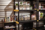 Several shelves are filled with various video game cases and boxes. The games are neatly organized, with some of them standing upright and others in stacks. The shelves are black with yellow accents and set against a metallic wall. There is a display box labeled 'Video Games', and various themed toys and merchandise are also visible.