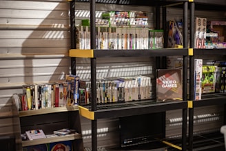 Several shelves are filled with various video game cases and boxes. The games are neatly organized, with some of them standing upright and others in stacks. The shelves are black with yellow accents and set against a metallic wall. There is a display box labeled 'Video Games', and various themed toys and merchandise are also visible.