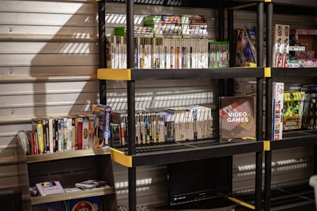 Several shelves are filled with various video game cases and boxes. The games are neatly organized, with some of them standing upright and others in stacks. The shelves are black with yellow accents and set against a metallic wall. There is a display box labeled 'Video Games', and various themed toys and merchandise are also visible.