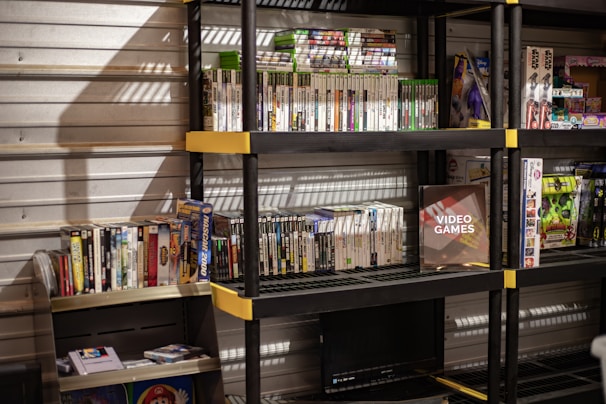 Several shelves are filled with various video game cases and boxes. The games are neatly organized, with some of them standing upright and others in stacks. The shelves are black with yellow accents and set against a metallic wall. There is a display box labeled 'Video Games', and various themed toys and merchandise are also visible.
