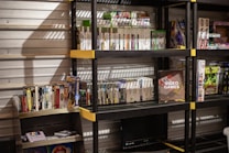 Several shelves are filled with various video game cases and boxes. The games are neatly organized, with some of them standing upright and others in stacks. The shelves are black with yellow accents and set against a metallic wall. There is a display box labeled 'Video Games', and various themed toys and merchandise are also visible.