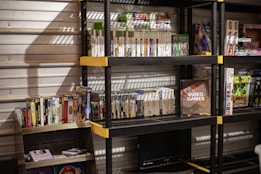 Several shelves are filled with various video game cases and boxes. The games are neatly organized, with some of them standing upright and others in stacks. The shelves are black with yellow accents and set against a metallic wall. There is a display box labeled 'Video Games', and various themed toys and merchandise are also visible.