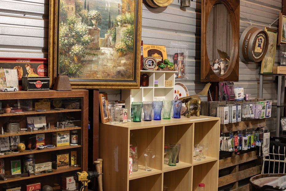 A vintage store display features a mix of old-fashioned objects and collectibles. There is a large framed painting of a scenic landscape, several collectible glasses in various colors on wooden shelves, and a variety of items such as vintage signs, mirrors, and decorative plates. Shelves are filled with retro memorabilia, including tins, books, and small model cars. The setting has a rustic and nostalgic atmosphere, with a mix of metal and wood materials.