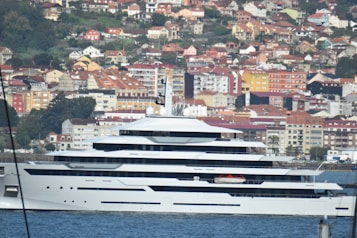 A large, modern yacht is docked in a harbor with a backdrop of densely packed houses and buildings on a hillside. The yacht is white with multiple decks and sleek lines. The background features various colorful buildings, with orange, red, and yellow rooftops, set amidst greenery.