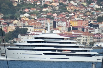 A large, modern yacht is docked in a harbor with a backdrop of densely packed houses and buildings on a hillside. The yacht is white with multiple decks and sleek lines. The background features various colorful buildings, with orange, red, and yellow rooftops, set amidst greenery.