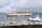 A large cargo ship loaded with multiple rows of orange containers navigates through a body of water. In the foreground, smaller boats are docked in a marina, and seagulls are flying above. The weather appears overcast, with clouds covering the sky and mountains in the background.