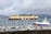 A large cargo ship loaded with multiple rows of orange containers navigates through a body of water. In the foreground, smaller boats are docked in a marina, and seagulls are flying above. The weather appears overcast, with clouds covering the sky and mountains in the background.