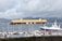 A large cargo ship loaded with multiple rows of orange containers navigates through a body of water. In the foreground, smaller boats are docked in a marina, and seagulls are flying above. The weather appears overcast, with clouds covering the sky and mountains in the background.