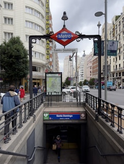 A city street features a subway entrance marked with a red and blue sign for 'Metro Santo Domingo'. The surrounding area includes tall buildings, visible traffic, and pedestrians. The skyline shows overcast skies with modern and historical architecture in the background.
