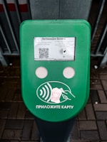 A green electronic device for ticket activation with instructions in Russian. It features a QR code, NFC symbol, and an illustration indicating where to place a card. The background consists of a pavement area partially visible.