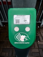 A green electronic device for ticket activation with instructions in Russian. It features a QR code, NFC symbol, and an illustration indicating where to place a card. The background consists of a pavement area partially visible.