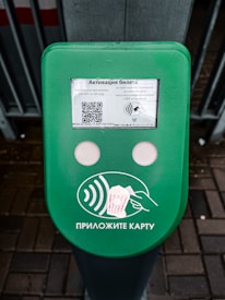 A green electronic device for ticket activation with instructions in Russian. It features a QR code, NFC symbol, and an illustration indicating where to place a card. The background consists of a pavement area partially visible.
