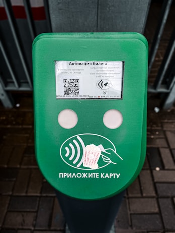 A green electronic device for ticket activation with instructions in Russian. It features a QR code, NFC symbol, and an illustration indicating where to place a card. The background consists of a pavement area partially visible.