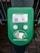 A green electronic device for ticket activation with instructions in Russian. It features a QR code, NFC symbol, and an illustration indicating where to place a card. The background consists of a pavement area partially visible.