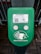 A green electronic device for ticket activation with instructions in Russian. It features a QR code, NFC symbol, and an illustration indicating where to place a card. The background consists of a pavement area partially visible.
