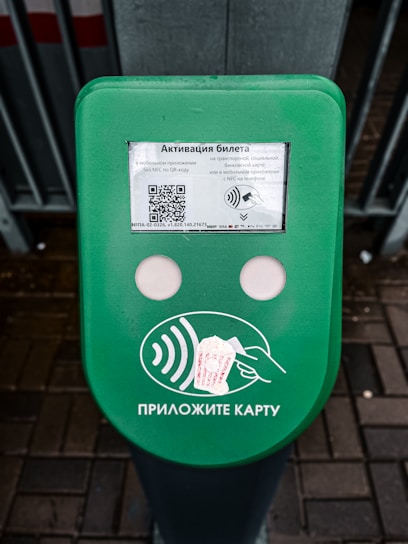 A green electronic device for ticket activation with instructions in Russian. It features a QR code, NFC symbol, and an illustration indicating where to place a card. The background consists of a pavement area partially visible.