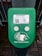 A green electronic device for ticket activation with instructions in Russian. It features a QR code, NFC symbol, and an illustration indicating where to place a card. The background consists of a pavement area partially visible.