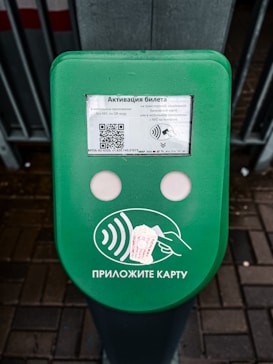A green electronic device for ticket activation with instructions in Russian. It features a QR code, NFC symbol, and an illustration indicating where to place a card. The background consists of a pavement area partially visible.