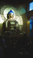 A golden statue of Buddha seated in a meditative pose. The statue is intricately detailed with a serene expression and a striking blue topknot. Behind the figure is an ornate circular backdrop with floral patterns. The ambient light gives a warm, sacred atmosphere. Flowers and small offerings are placed in front of the statue, adding to the spiritual ambiance.