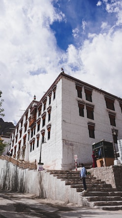 A large, traditional Tibetan building with whitewashed walls adorned with ornate window frames. The structure is located at the top of a set of stone steps and surrounded by a mountainous landscape. A person stands on the steps leading upwards, and the sky above is partly cloudy with patches of bright blue.
