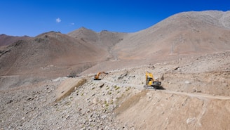 Heavy machinery operates on a rugged mountain landscape. Two yellow excavators are working on a dirt road that cuts through rocky terrain under a clear blue sky.